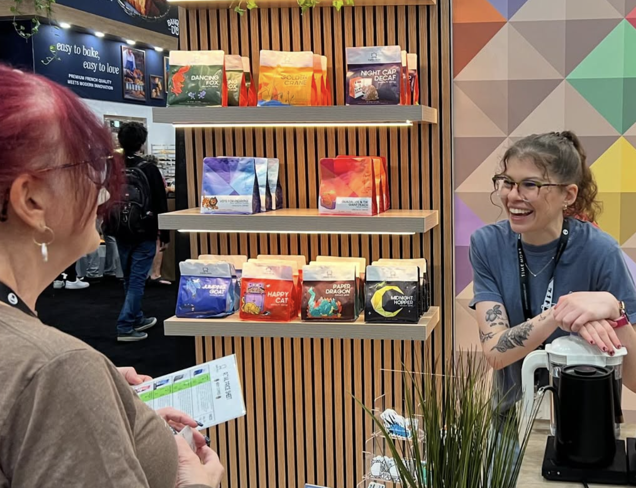 An excitable barista chats with a guest at a coffee booth in a convention center. Three shelves with colorful coffee bags are on the wall in the background.