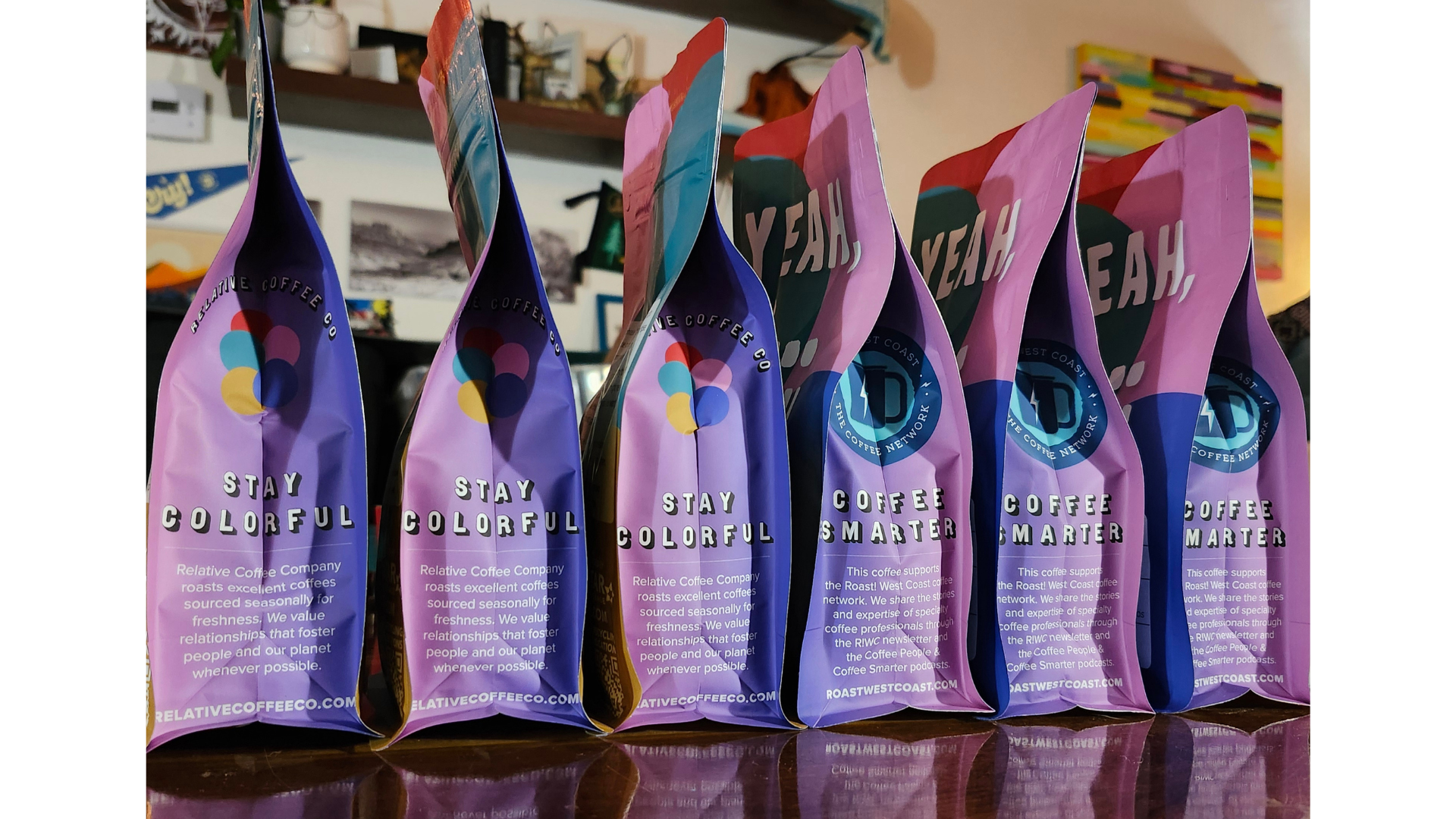 A close-up on six colorful coffee packages side-by-side. The first three say "Stay Colorful" with copy below. The second three say "Coffee Smarter" with copy below. Each bag is reflected slightly in the gloss of a wooden table top at the forefront of the image.