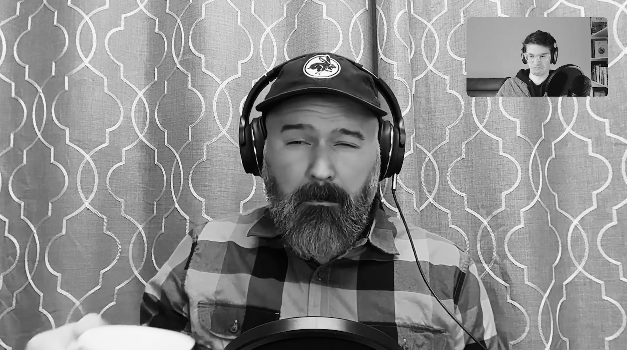 A bearded man in a cap, plaid print shirt, and over the ear headphones makes a scrunched face at the camera after taking a sip of coffee.