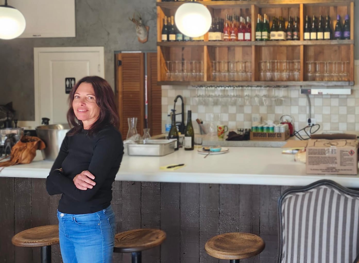 A caucasian woman in a black top and jeans folds her arms and smiles at the camera in front of a wine bar that is under construction.