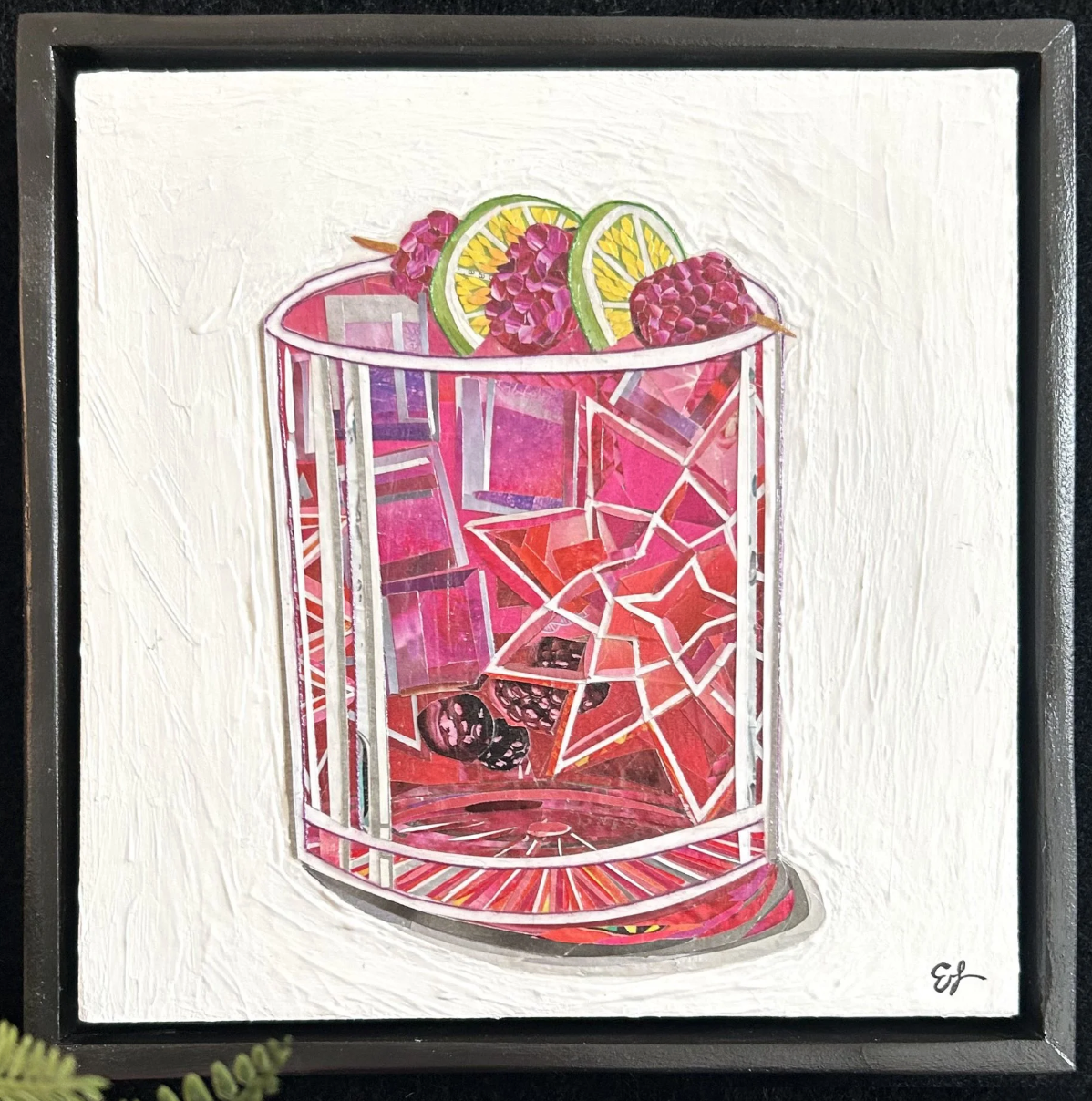 A close up of a framed paper collage art-piece featuring cut paper overlapped to create a red-colored cocktail with bright green lime slices against a white background.