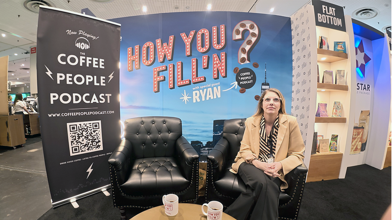 A caucasian woman with long blond hair, glasses, wearing a tan overcoat and black slacks sits cross-legged in a comfy black faux leather lounge chair in front of a convention booth backdrop made to mimic a late-night studio.